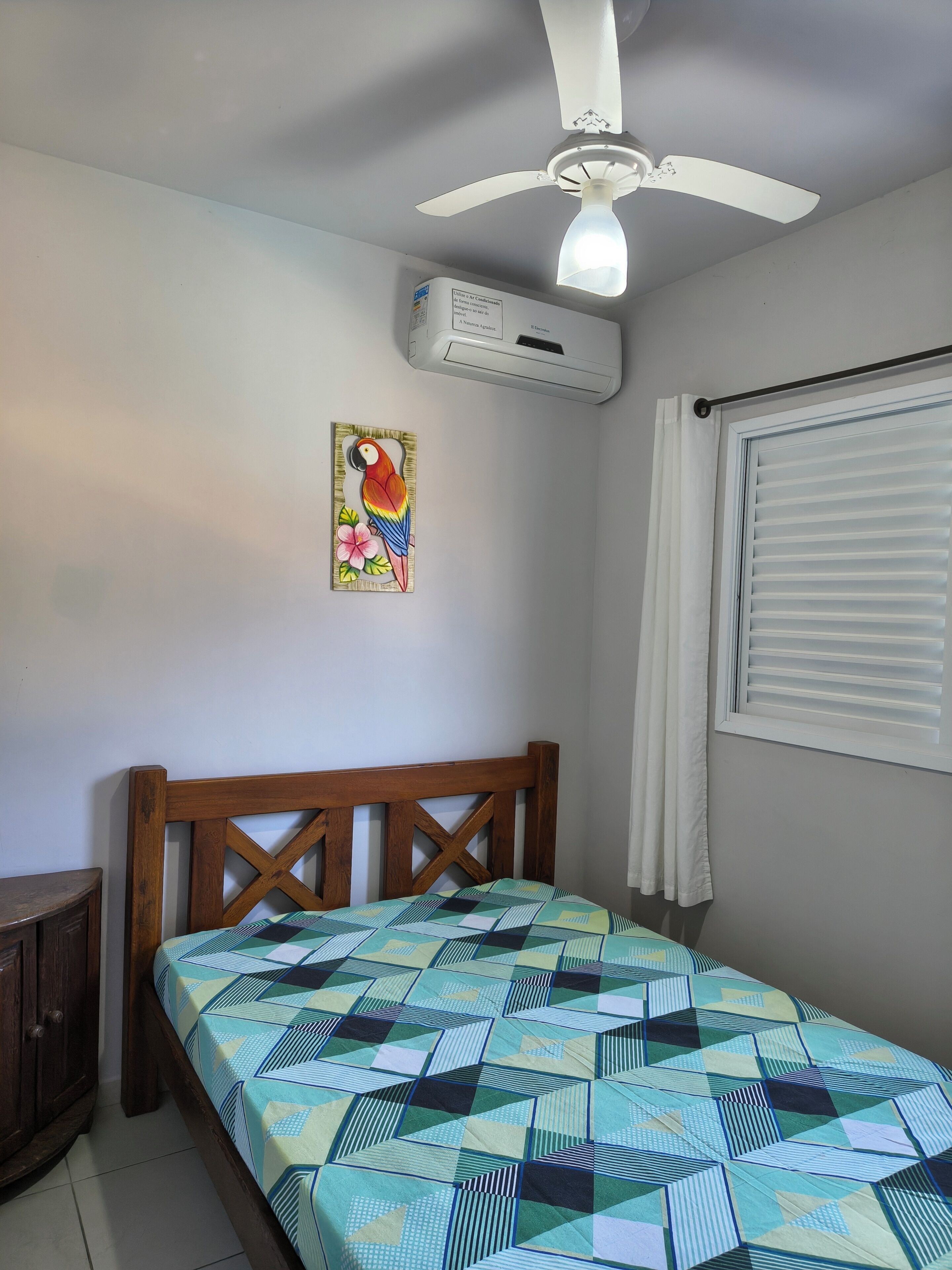 Ein Schlafzimmer mit einem hölzernen Bettgestell, einer Bettdecke, einem Deckenventilator, einer Klimaanlage, einem Fenster mit Jalousien und einer Wand-Klimaanlage.