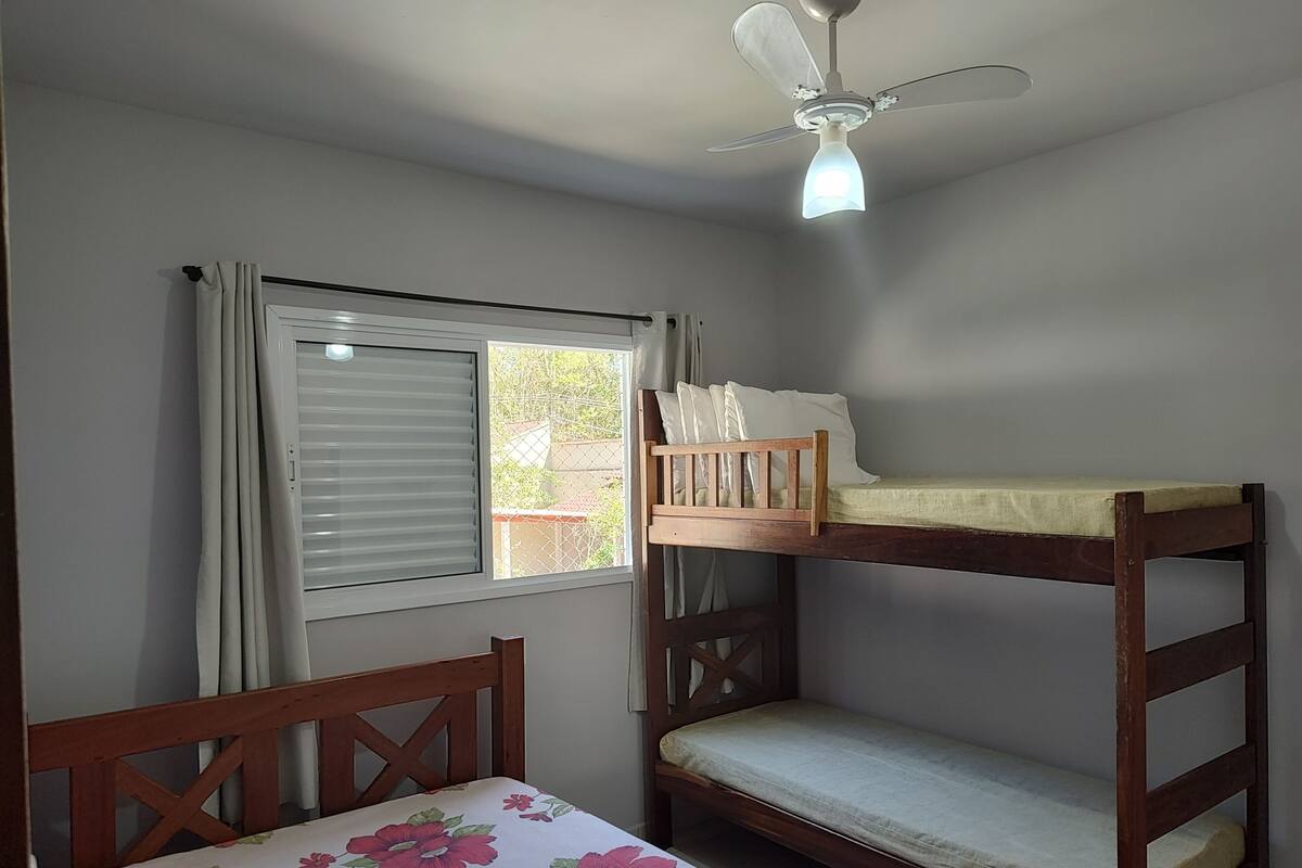 A bunk bed with a wooden headboard and a single bed with a floral bedspread. A ceiling fan is visible above the bed, and a window with curtains is present.