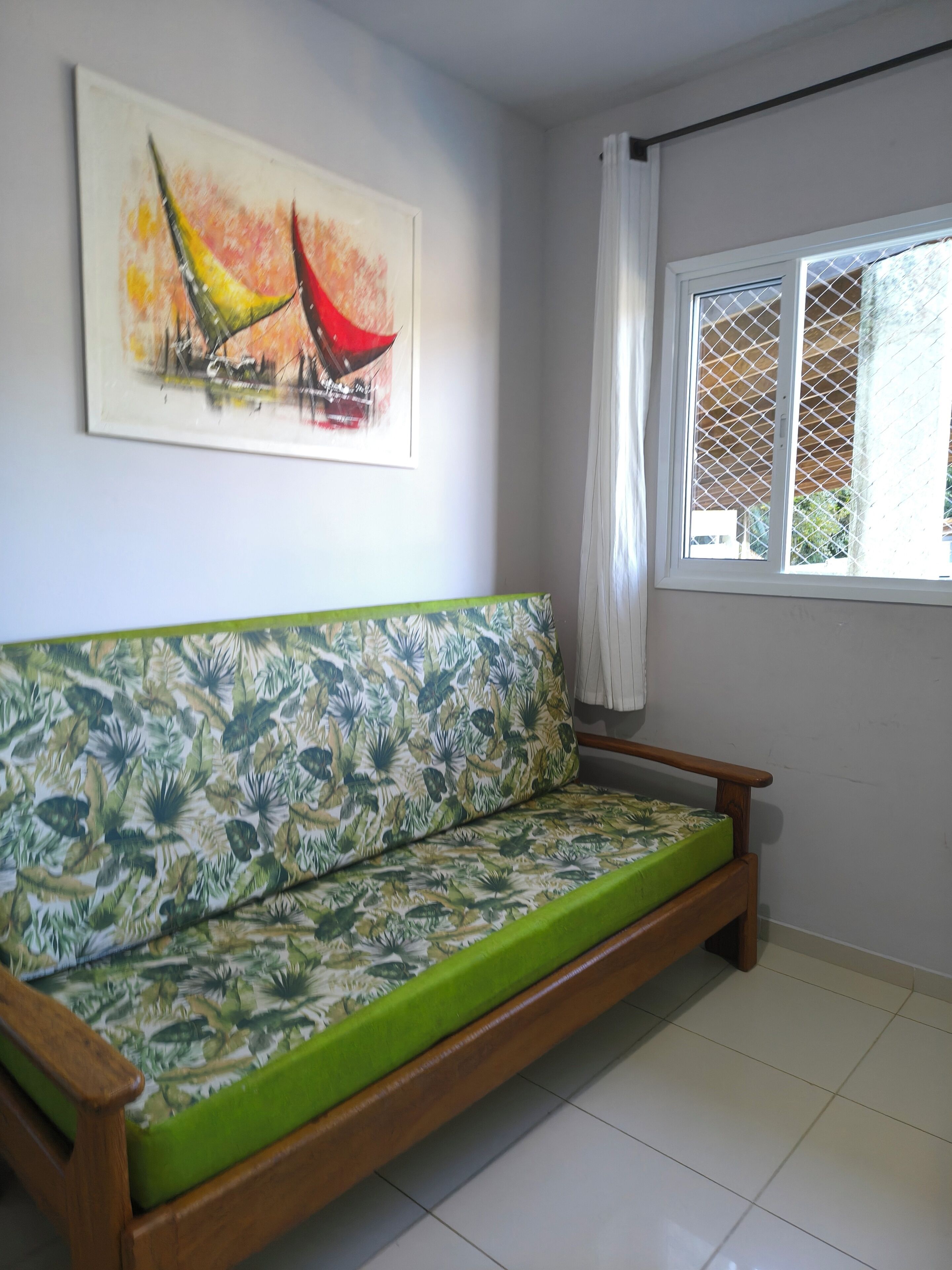 Ein Zimmer mit einer Holzsofa, einem grünen Kissen, einem Bild an der Wand und einem Fenster mit weißem Vorhang.