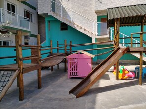 A playground with wooden climbing structures, a pink slide, and colorful play equipment. - Canto Verde Apartamentos Caraguá (Caraguatatuba)