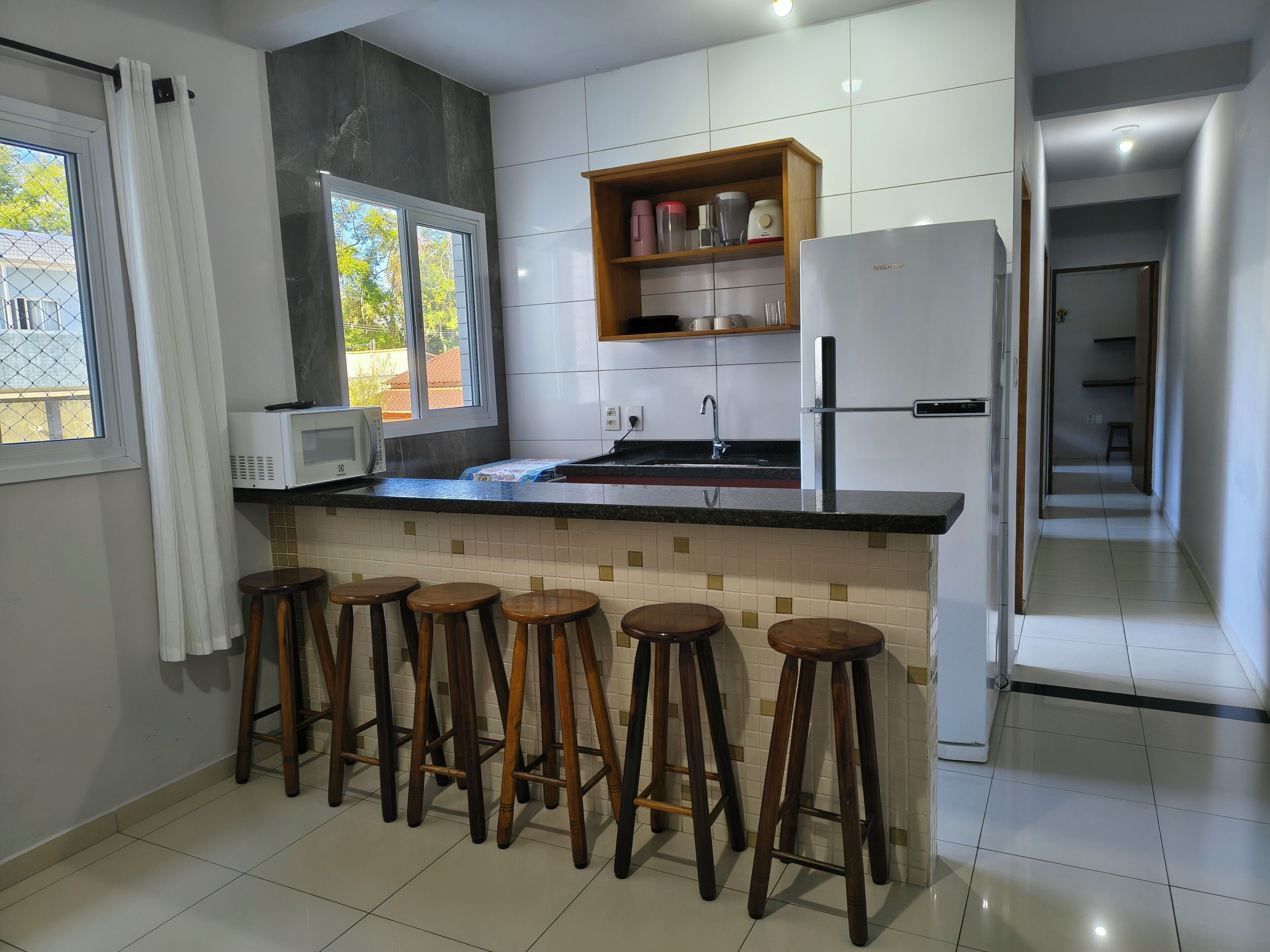 A modern kitchen with a white refrigerator, a microwave, and a wooden shelf with kitchenware. There are four wooden stools under a window.
