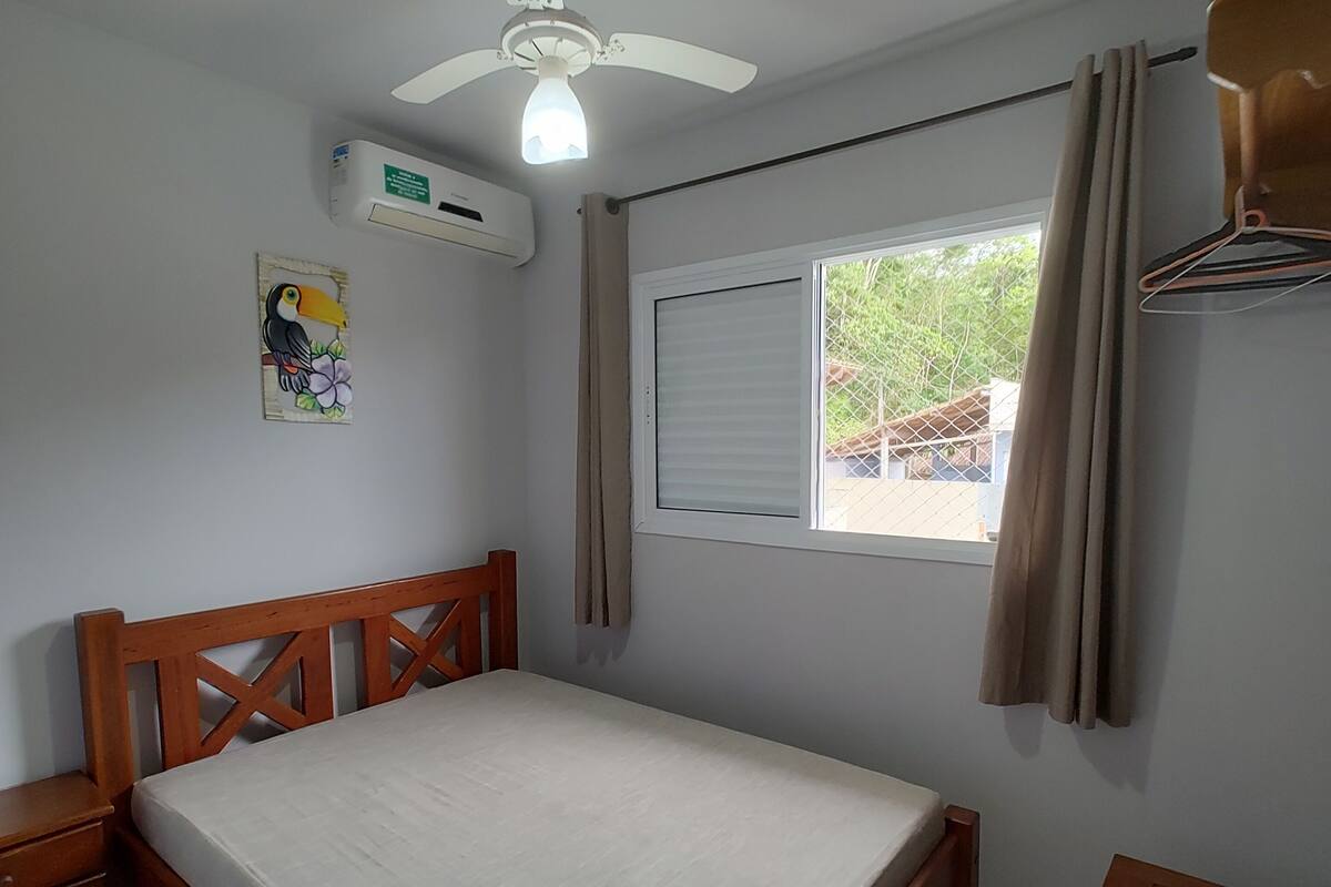 A bedroom with a wooden bed, a ceiling fan, an air conditioning unit, a window with curtains, and a small table.