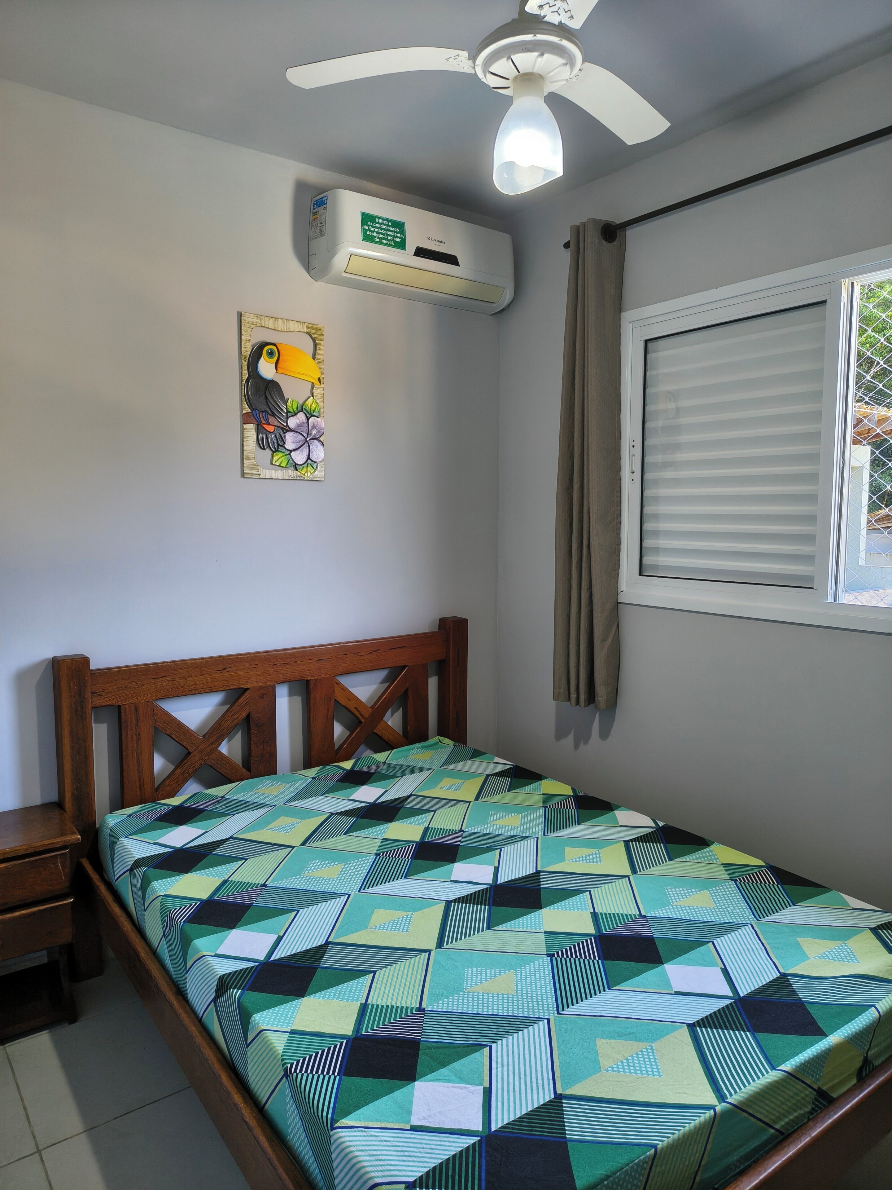 A bedroom with a bed, a ceiling fan, an air conditioning unit, a window with curtains, and a wall-mounted picture.