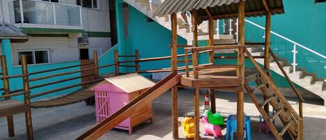 A playground with a wooden slide and climbing structures, surrounded by a building with a green exterior and multiple windows.