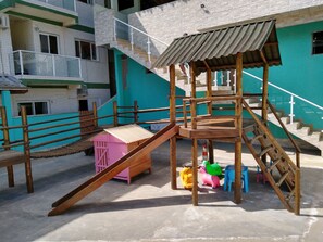 A playground with a wooden slide and climbing structures, surrounded by a building with a green exterior and multiple windows. - Canto Verde Apartamentos Caraguá (Caraguatatuba)