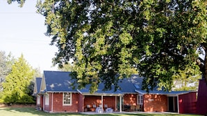 A brick building with a covered patio area, a table with chairs, and a large tree in front. - Coldstream Estate - The Whare (Coldstream)