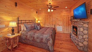 A cozy cabin room with a wooden ceiling, a bed with patterned bedding, a stone fireplace, and a flat-screen TV mounted above it. - Moonstruck Apartment 1 (Gatlinburg)