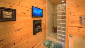 A wooden cabin bathroom with a fireplace, a television, and a glass shower enclosure. - Moonstruck Apartment 1 (Gatlinburg)