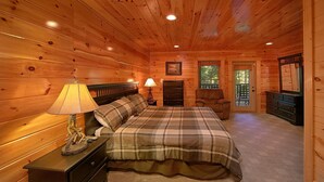 A cozy log cabin bedroom with a bed, two bedside lamps, a dresser, a chair, and a door leading outside. - Natural Attractions Apartment 3 (Gatlinburg)