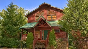 A log cabin with a green roof, wooden porch, and a stone wall. - Natural Attractions Apartment 3 (Gatlinburg)
