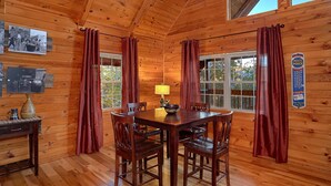 A wooden cabin interior with a dining table, chairs, a lamp, and two windows with curtains. - Moonshiners View Apartment 2 (Gatlinburg)