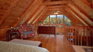 A cozy attic bedroom with wooden beams, a floral sofa, a dresser, and a large window offering a scenic view. - Moonshiners View Apartment 2 (Gatlinburg)