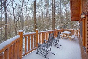 Une terrasse en bois, agrémentée de chaises et d’une table en métal, entourée d’une balustrade en bois, le tout se dressant sur fond de forêt enneigée.