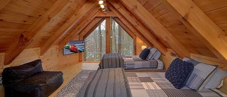 Ein Schlafzimmer im Dachgeschoss mit schrägen Wänden, Holzbalken, einem großen Fenster, einem Bett, einer Couch und einem Fernseher.