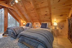 Une chambre confortable située sous les toits, avec deux lits, des murs en bois et une grande fenêtre donnant sur la forêt environnante.