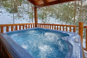 Un bain à remous sur une terrasse en bois, avec une forêt enneigée en arrière-plan.
