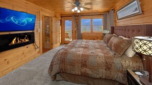A cozy log cabin bedroom with a fireplace, a large bed with patterned bedding, a ceiling fan, and a view of the mountains through the windows.