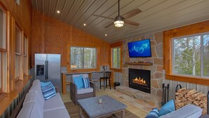 A cozy living room with a stone fireplace, a television, and a large window overlooking the forest.