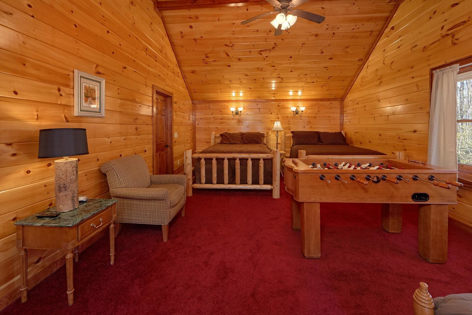 A cozy room with wooden walls, a ceiling fan, a bed, a sofa, a table with a lamp, and a foosball table.