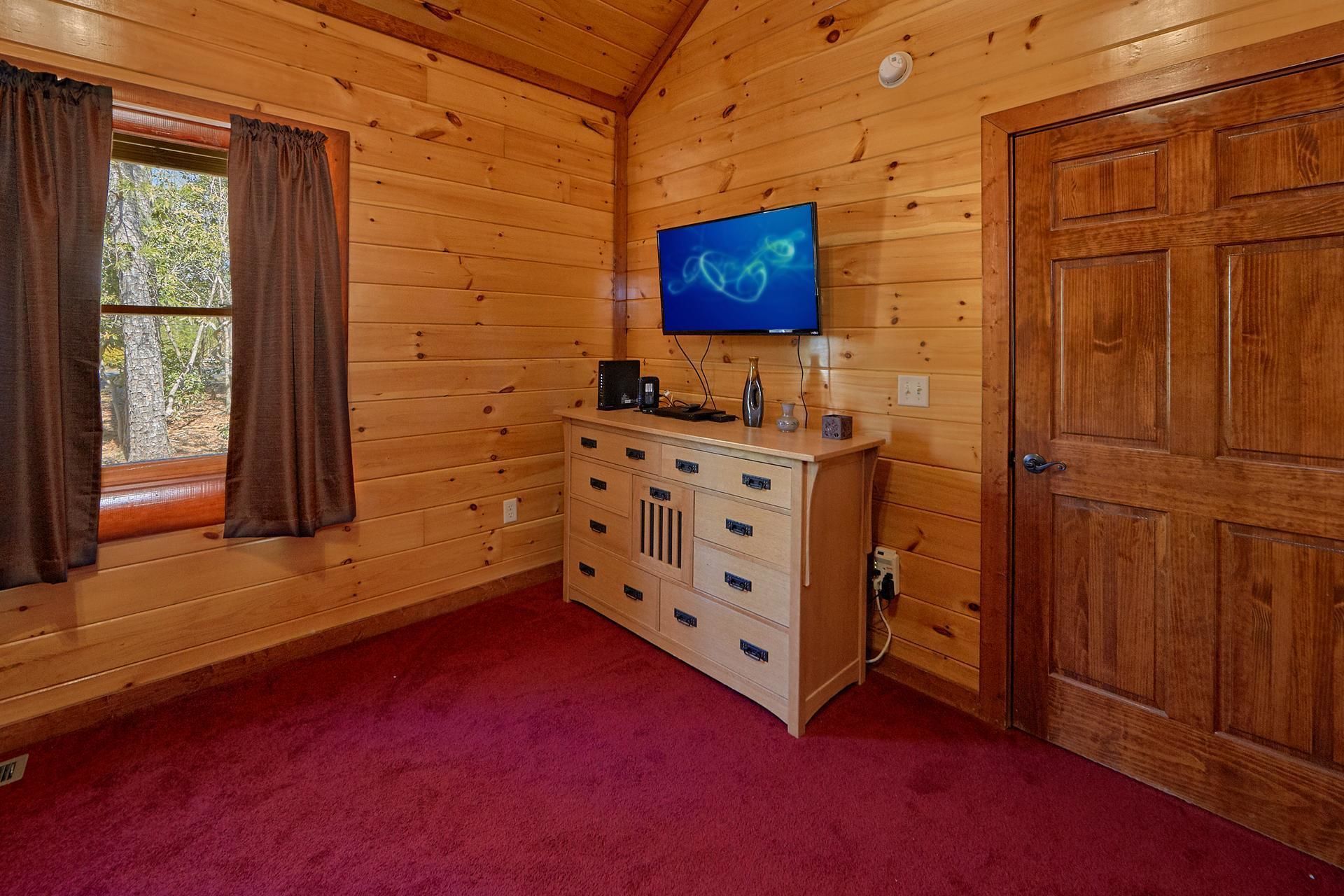 A wooden-paneled room with a television mounted on the wall, a wooden cabinet with drawers, a door, and a window with curtains.