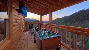 A wooden deck with a foosball table and a TV mounted on the wall, overlooking a forested mountain range.
