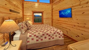A cozy wooden cabin room with a bed, a nightstand, a lamp, a TV, and a window with a scenic view.