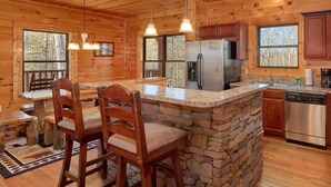 Ein Interieur einer Hütte mit einer steinernen Bar im Inselstil, Holzstühlen, einem Kühlschrank und einer Kaffeemaschine.