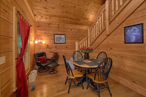 Interior de una cabaña de madera con comedor, escalera y ventana con cortinas rojas.