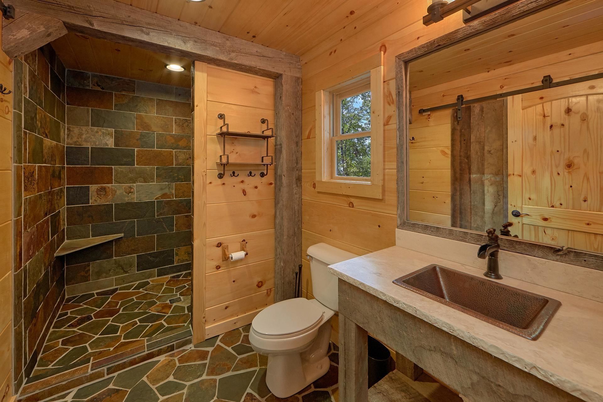 Une salle de bain en bois avec une douche carrelée en pierre, des toilettes, un lavabo et une fenêtre.