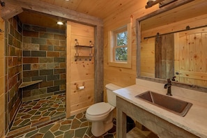 A wooden bathroom with a stone-tiled shower, a toilet, a sink, and a window.