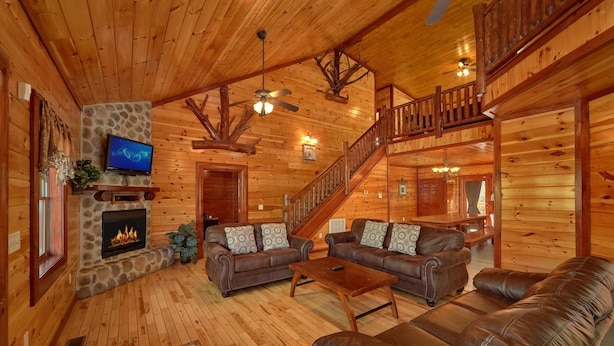 A log cabin interior with a fireplace, leather sofas, a TV, and a staircase. - Pool Movie 1 Apartment 6 (Cosby)