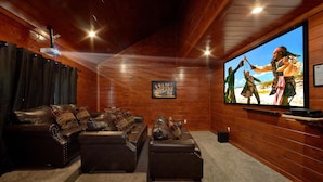 A cozy room with a leather couch, ottoman, and a large TV screen displaying a movie scene. - Pool Movie 1 Apartment 6 (Cosby)