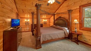 A four-poster bed in a log cabin room with a ceiling fan, a television on a wooden dresser, and a window with a view of trees. - Pool Movie 1 Apartment 6 (Cosby)
