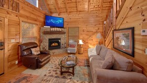 Interior acogedor de una cabaña de troncos con chimenea, sillón de cuero, mesa de centro de vidrio y televisor montado en la pared.