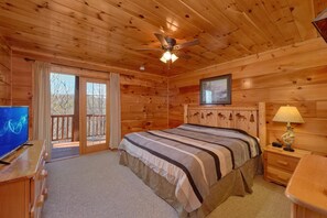 A wooden cabin bedroom with a bed, a television, a ceiling fan, and a window with a view of trees.