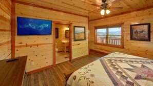 A wooden cabin bedroom with a bed, a ceiling fan, a TV, a bathroom, and a window with a mountain view. - Summit Pool Lodge Apartment 6 (Gatlinburg)
