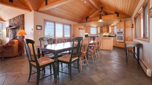 Un vaste espace de salle à manger avec une grande table et des chaises en bois, une cuisine dotée de meubles et d’appareils électroménagers en bois, et une cheminée en pierre.