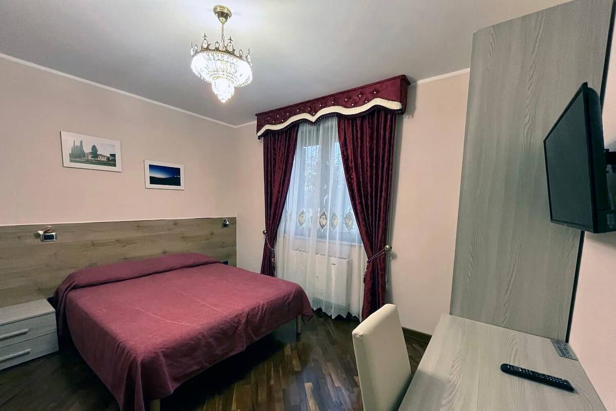 A hotel room with a bed, a desk, a TV, a chandelier, and a window with curtains.