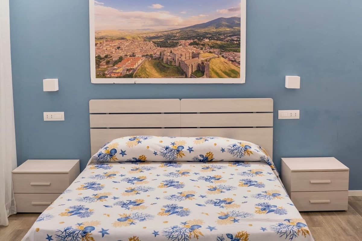 A bedroom with a bed, two bedside tables, a nightstand, a wall-mounted picture, and a light switch.