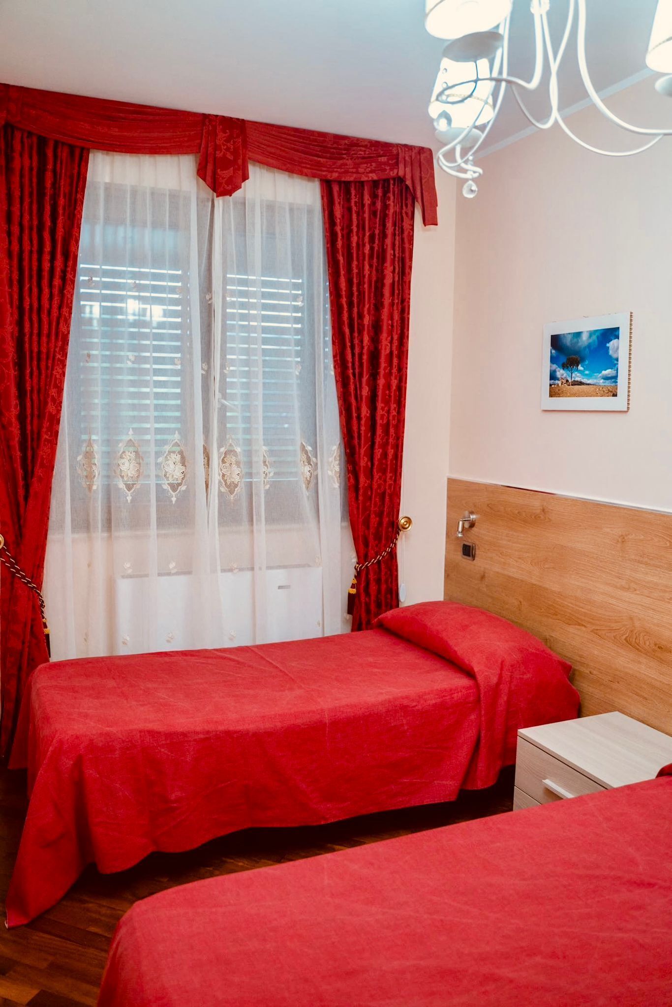 A hotel room with two beds, red bedding, a wooden headboard, a white nightstand, and a window with red curtains.