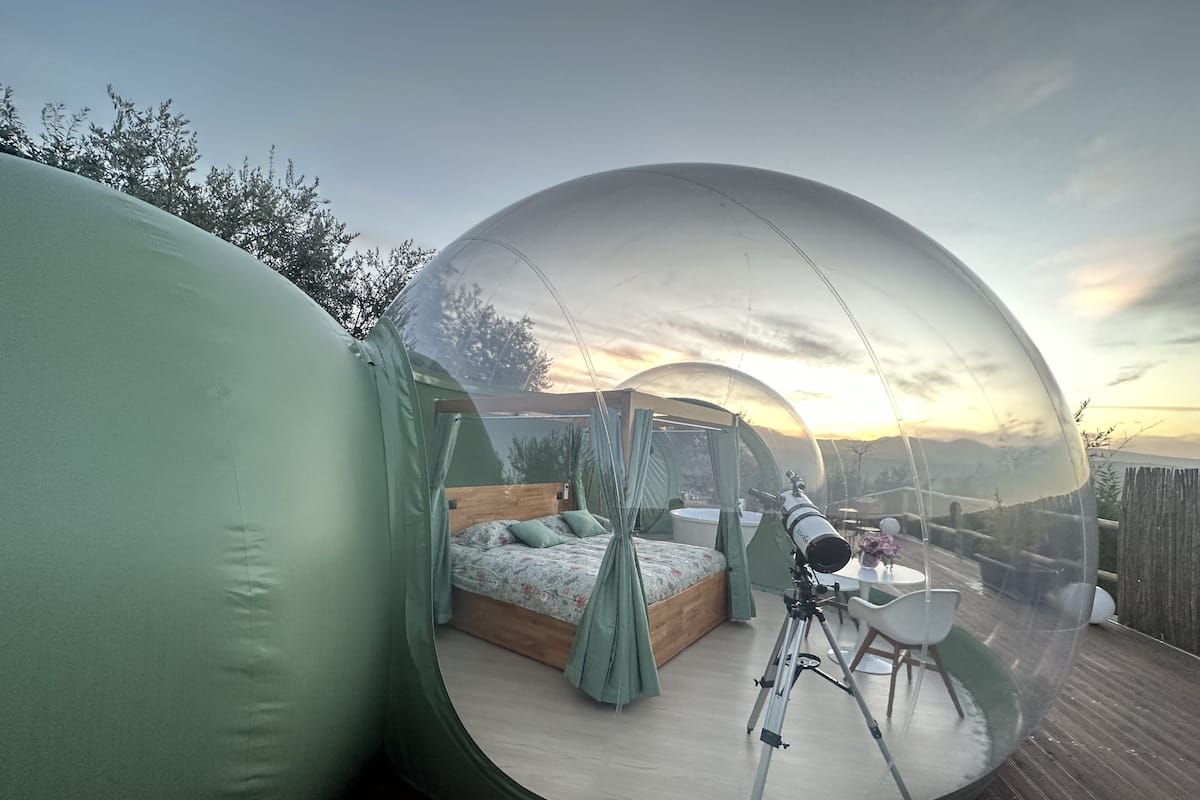 A transparent dome room with a bed, a telescope, and a view of the sunset.