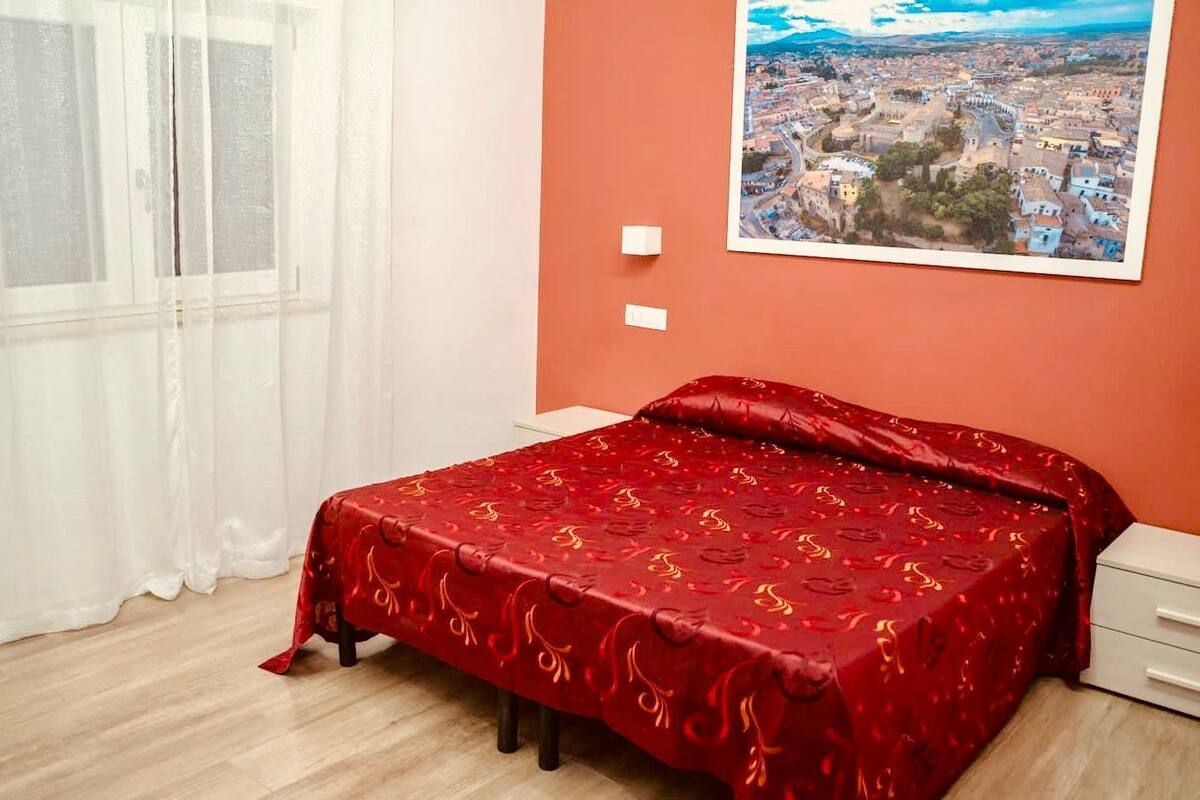 A bedroom with a bed, a window with white curtains, a white nightstand, and a framed cityscape picture on the wall.