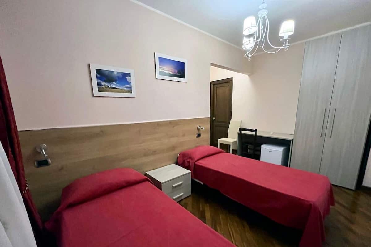 A hotel room with two single beds, a desk, a chair, a wardrobe, and two wall-mounted pictures.