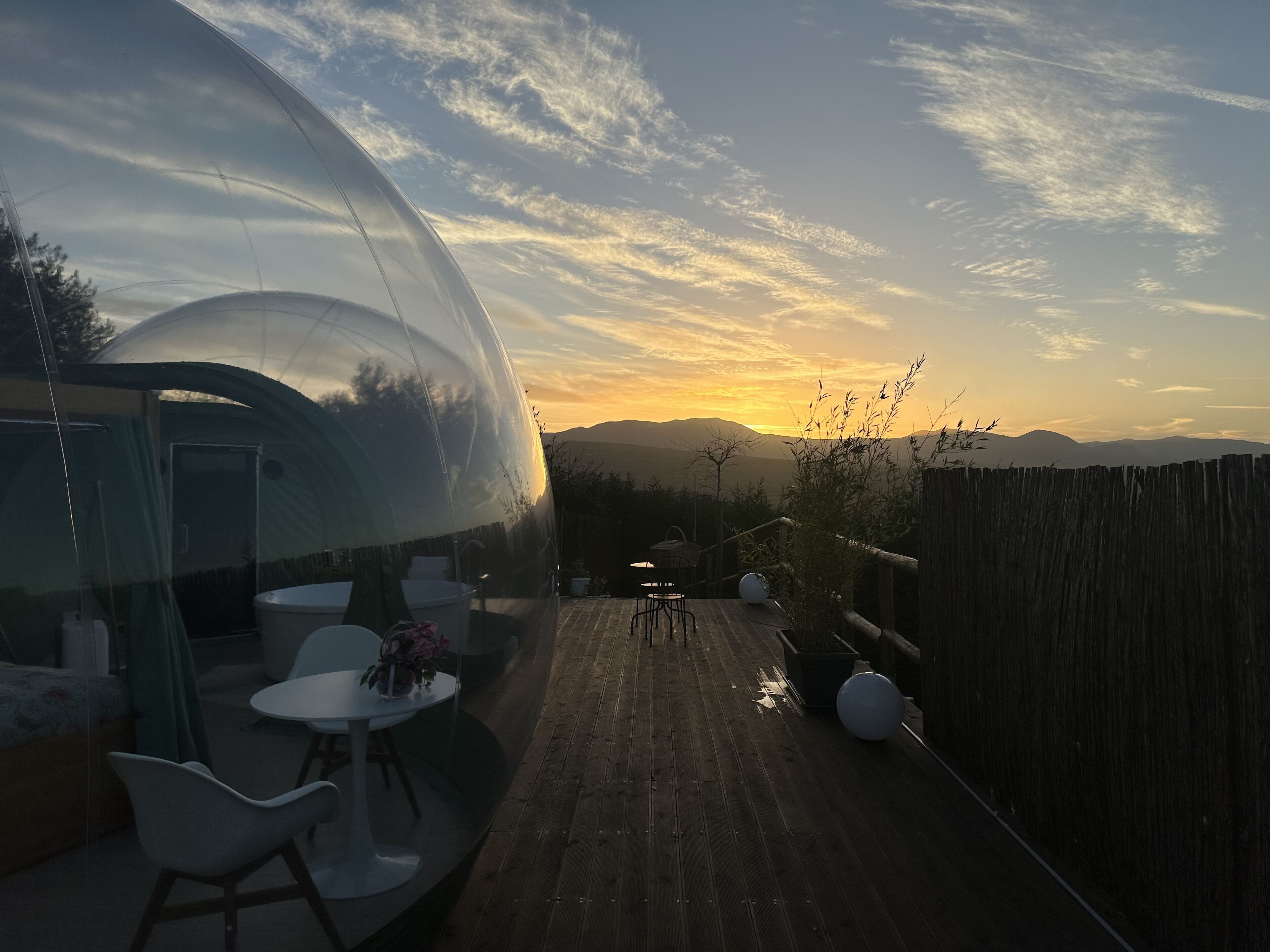 Una estructura de cúpula transparente con una mesa y sillas en su interior, con vistas a una terraza de madera y montañas al fondo.