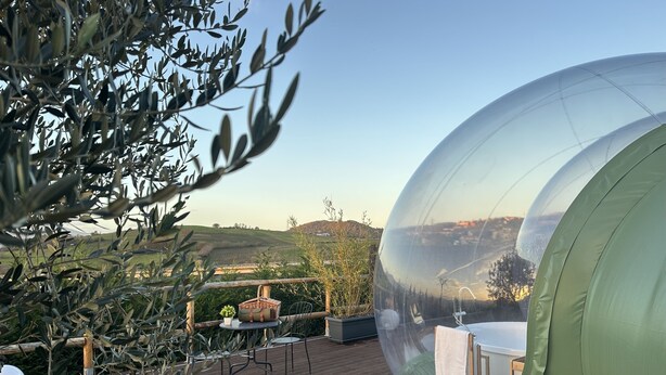 Estructura abovedada transparente con terraza de madera, asientos al aire libre y vista a un paisaje montañoso.