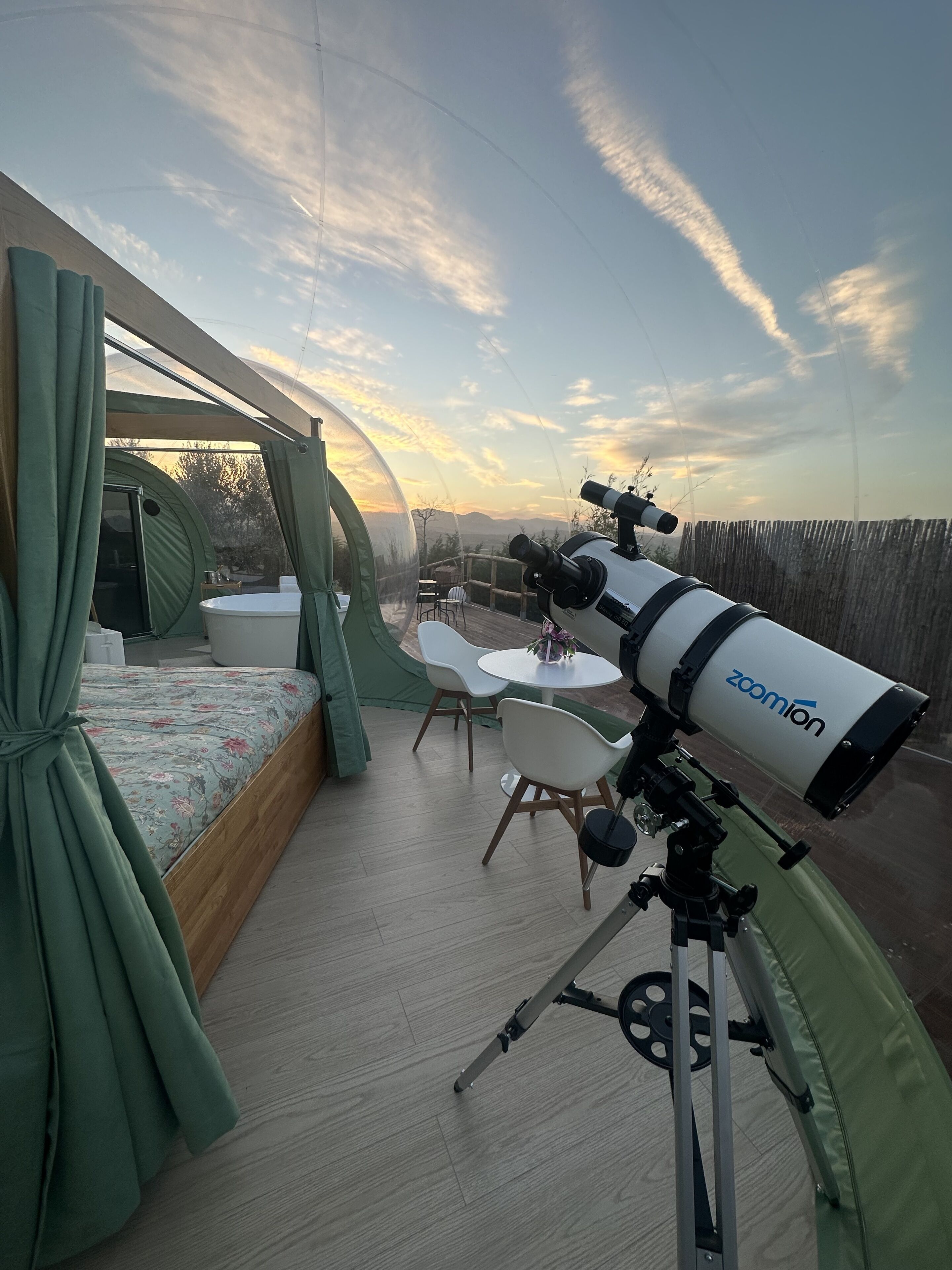 Un telescopio su un treppiede, un letto con tende verdi, un tavolo rotondo e sedie in una stanza con vista sul cielo.