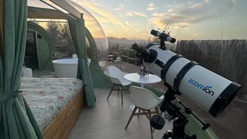 Un telescopio su un treppiede, un letto con tende verdi, un tavolo rotondo e sedie in una stanza con vista sul cielo.