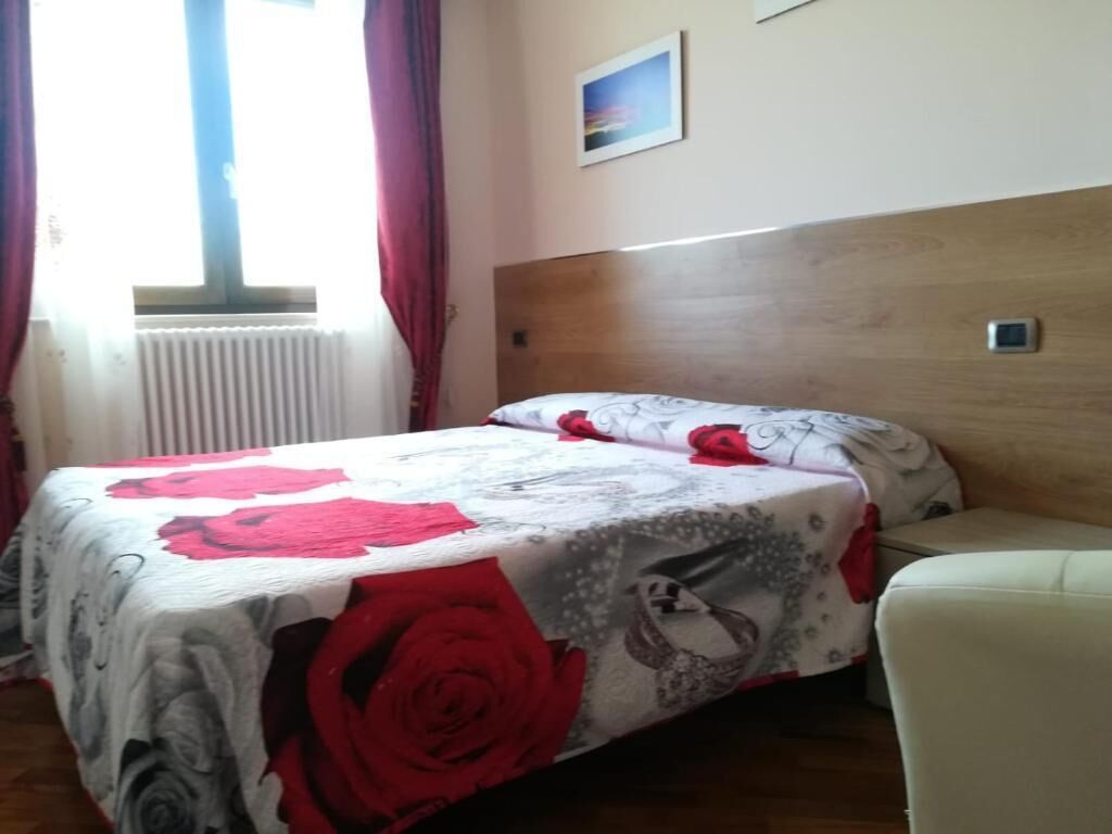 A bedroom with a bed, a wooden headboard, a window with curtains, a radiator, and a small painting on the wall.