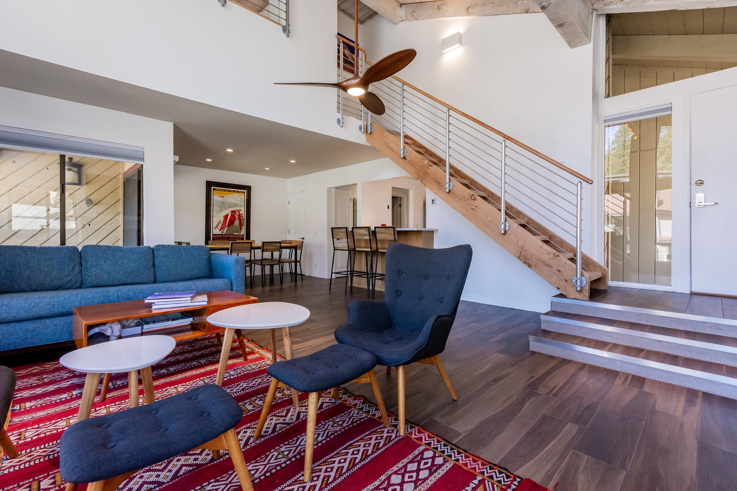 A spacious living area with a blue sofa, two armchairs, a coffee table, and a patterned rug. A wooden staircase with a metal railing leads to an upper floor.