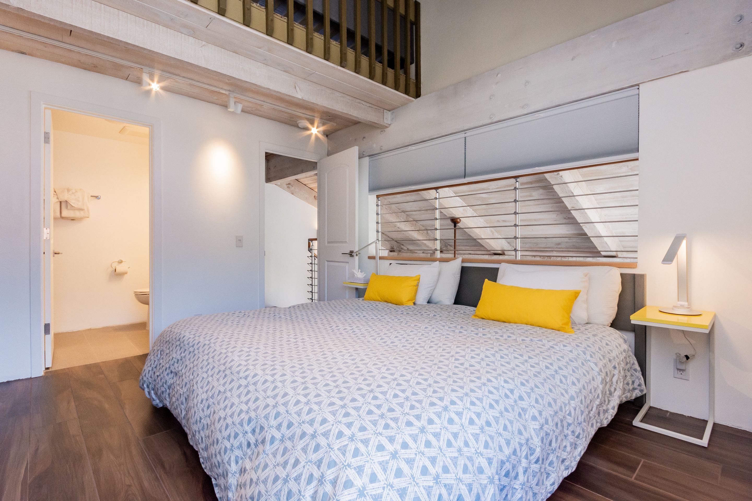 Une chambre avec un grand lit, un plancher en bois, un puits de lumière et une petite table de chevet avec une lampe.